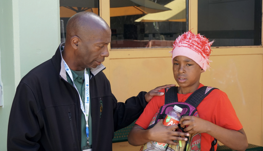A Black man wearing a black jacket rests his left hand on a young Black boy's shoulder who is earring a red hat, red shirt and a backpack on his chest while holding a beverage.